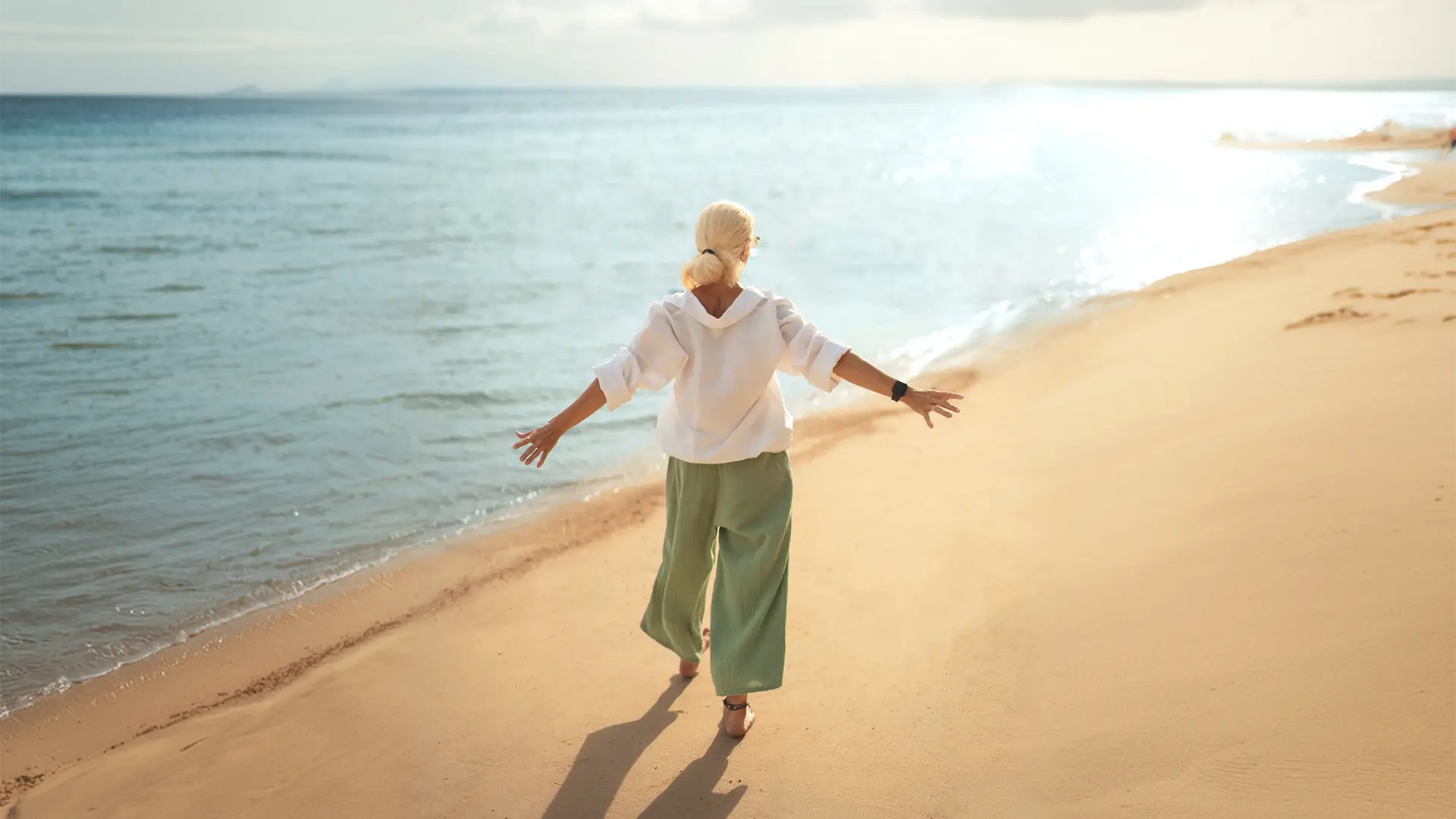 Donna che cammina sulla spiaggia con le braccia aperte, che simboleggia l'energia e il benessere supportati dall'idrogeno molecolare transdermico.